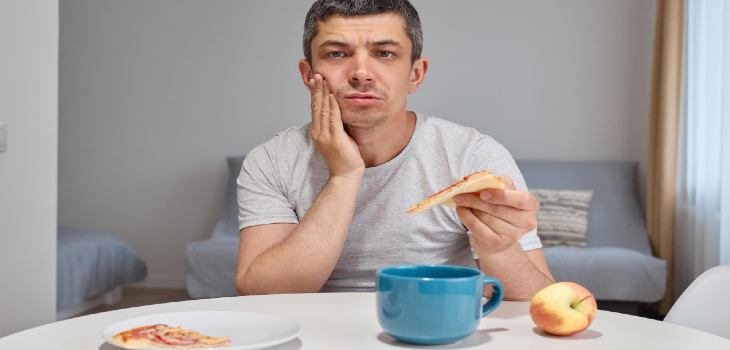 Disappointed upset young man holding piece of pizza feeling teeth pain after eating junk food touching his painful cheek